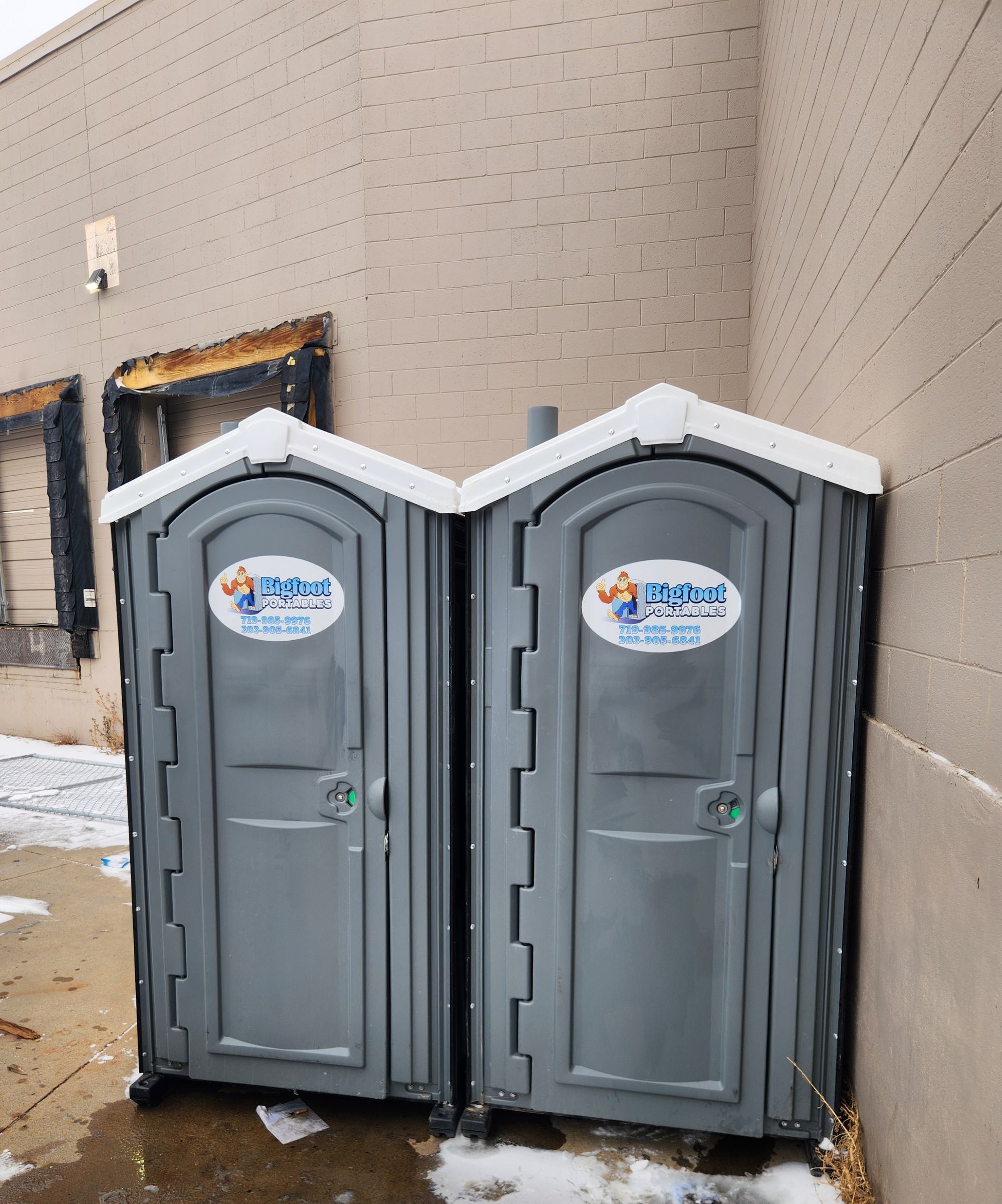 Two Bigfoot Portables porta potties placed beside a commercial building